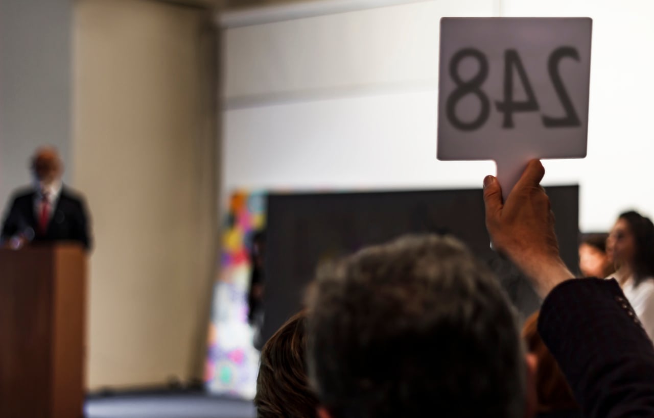 A person holds up a bidding paddle with the number 248 at an auction, while an auctioneer stands in the background at a podium.