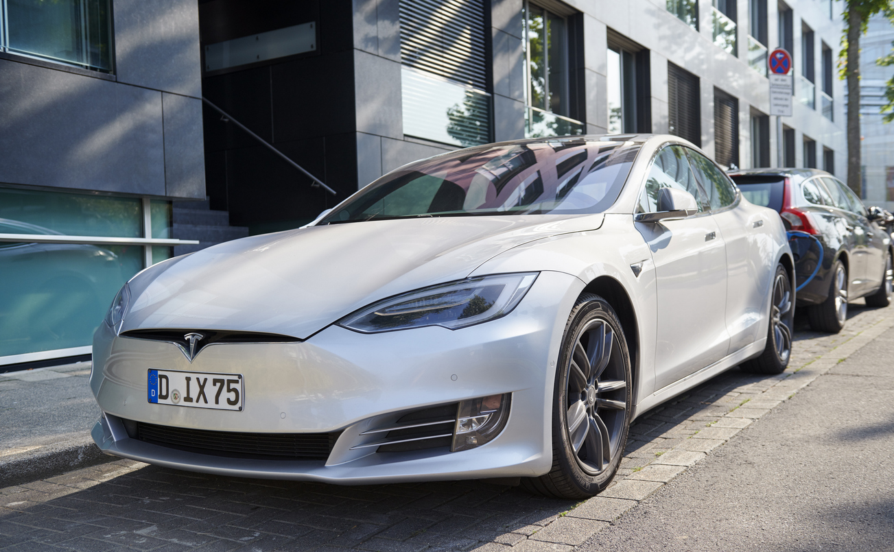 A silver Tesla Model S electric car is parked on a city street beside a modern glass building. Other vehicles are parked behind it, and trees reflect on the car's windows.