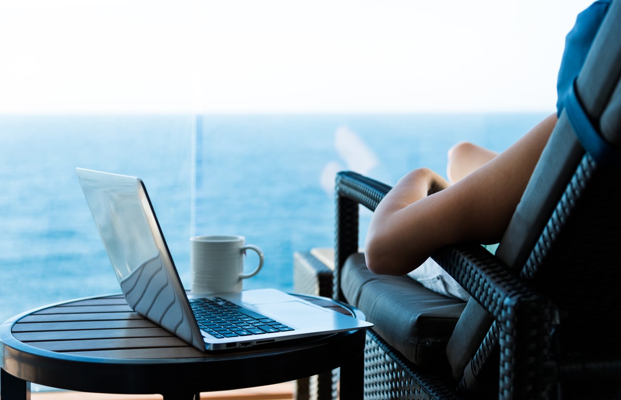 A person relaxes on a lounge chair facing the ocean, with a laptop and a coffee mug placed on a small table beside them. The scene is calm, bright, and suggests remote work or leisure by the sea.