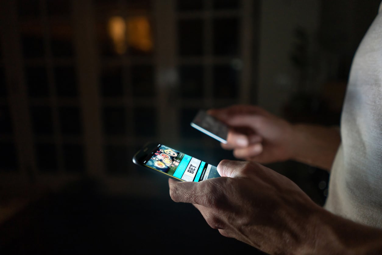 A person stands indoors in dim lighting, holding a smartphone and a credit card. The phone screen displays a food delivery app with images of various dishes. The person's face is not visible.