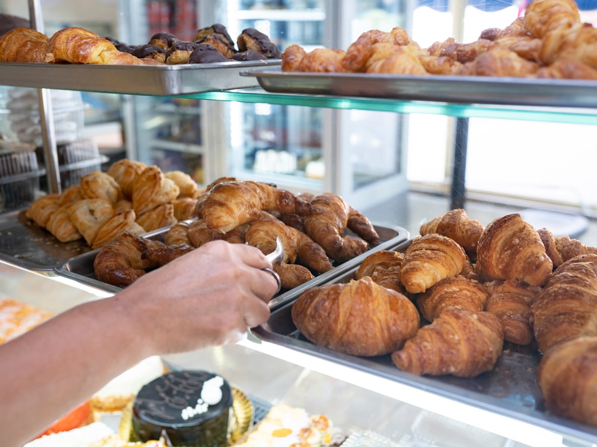 A person’s hand uses tongs to pick up a croissant from a display case filled with fresh croissants and pastries in a bakery. Other pastries are visible on the shelves and behind the glass.