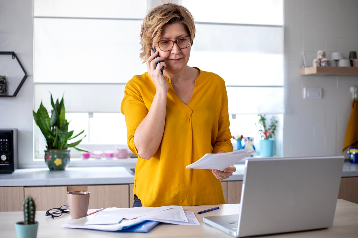 A woman in a yellow shirt stands in a bright kitchen, talking on the phone while looking at documents. An open laptop, papers, a mug, and a small plant are on the table in front of her.