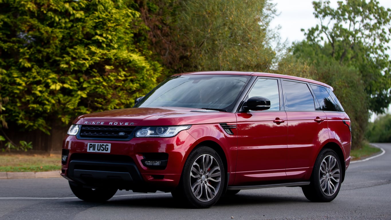 A red Range Rover SUV is parked on a suburban street, surrounded by green trees and bushes on a sunny day.