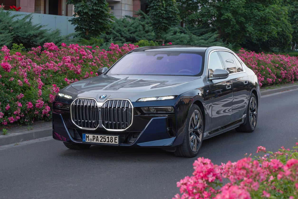 A dark-colored BMW sedan with a prominent front grille is parked on a street lined with blooming pink flowers and green bushes.