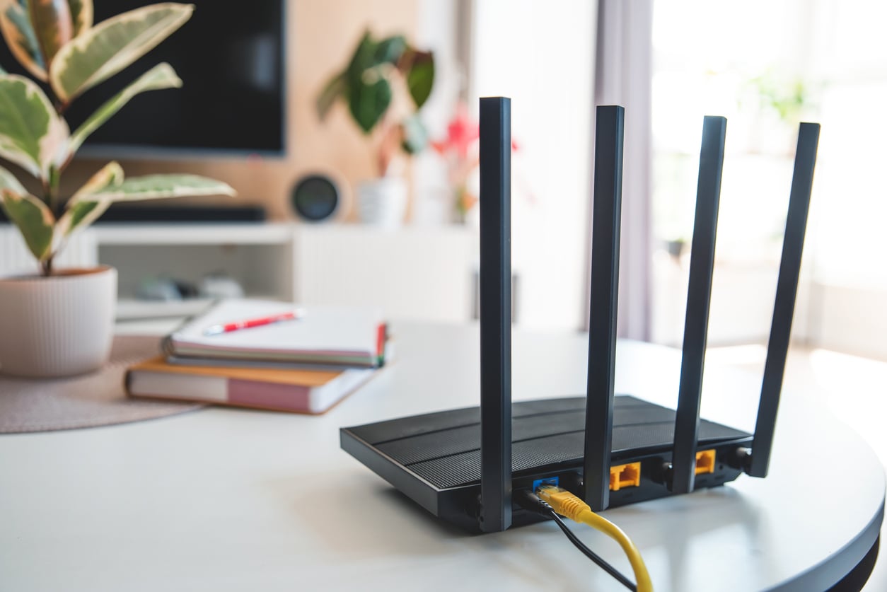 A wireless Wi-Fi router with four antennas sits on a white table. In the background are houseplants, books, a pen, and a blurred TV, suggesting a cozy and modern home environment.