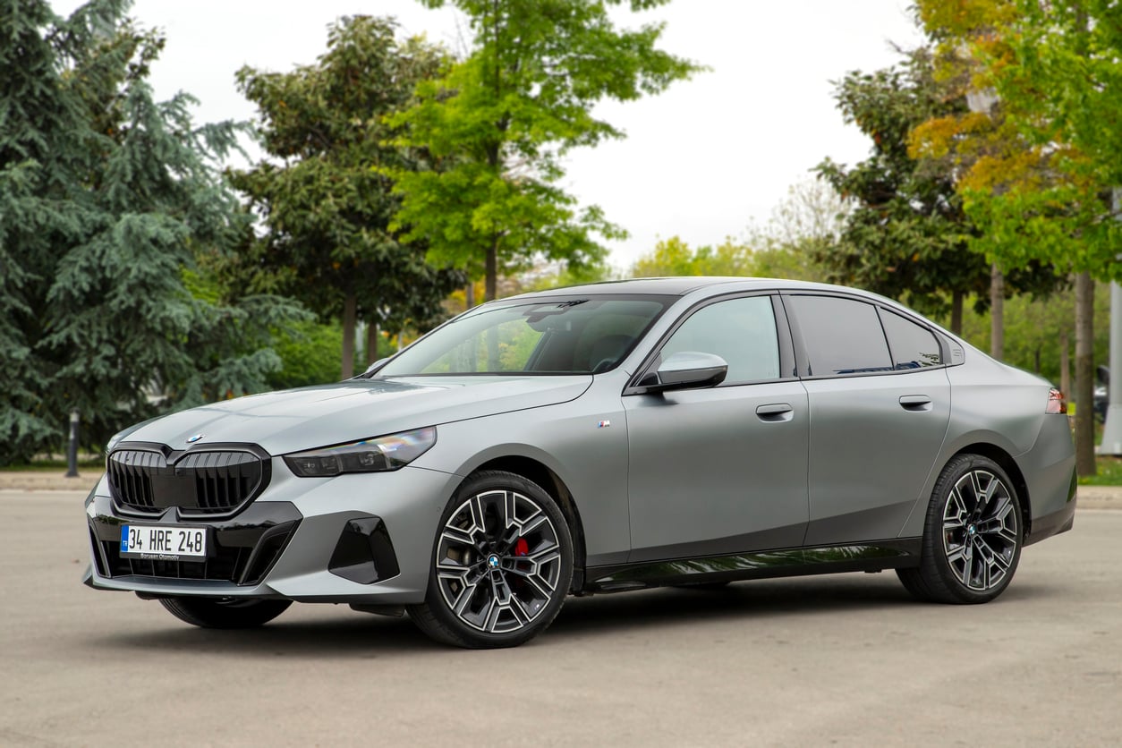 A silver BMW sedan is parked on a paved road, surrounded by green trees and a cloudy sky in the background. The car features sporty black and silver wheels and tinted windows.