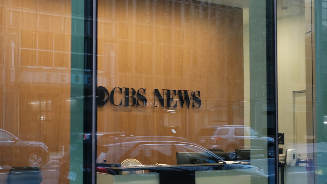 A glass window displays the CBS News logo on a wooden wall inside an office, reflecting cars and buildings from the street—a quiet scene amid ongoing talks about potential 2026 layoffs.