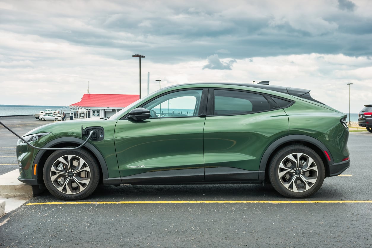 A green electric SUV is parked and charging at a station in a parking lot near a building with a red roof, with a cloudy sky in the background.