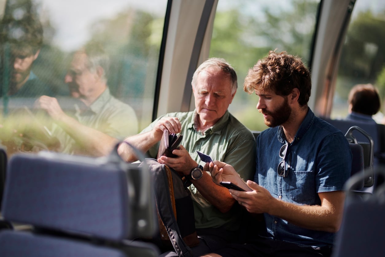 Two men sit side by side on a train, one older and one younger, both looking at a smartphone together. Sunlight streams through the window, and their reflections are visible in the glass.