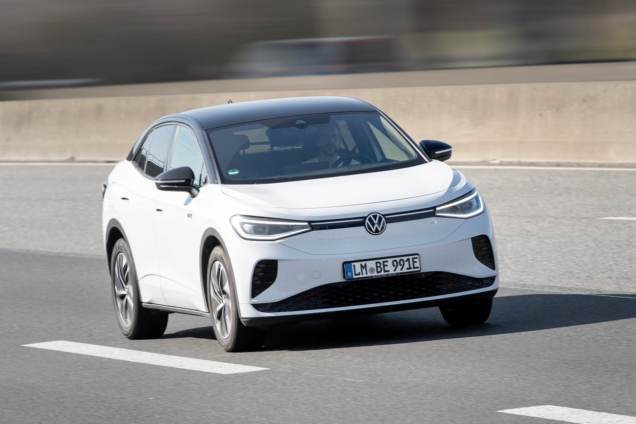 A white Volkswagen electric SUV with a black roof is driving on a multi-lane road. The driver is visible inside the vehicle. The background appears blurred, indicating motion.