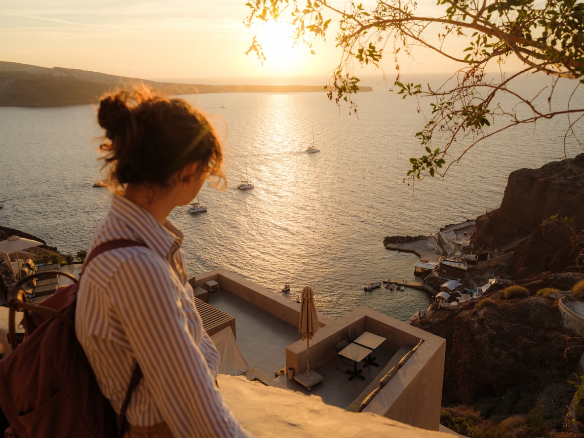 A woman with a backpack gazes at the sunset over the sea from a terrace on a cliff, with boats on the water and coastal buildings below in a scenic, warm landscape.