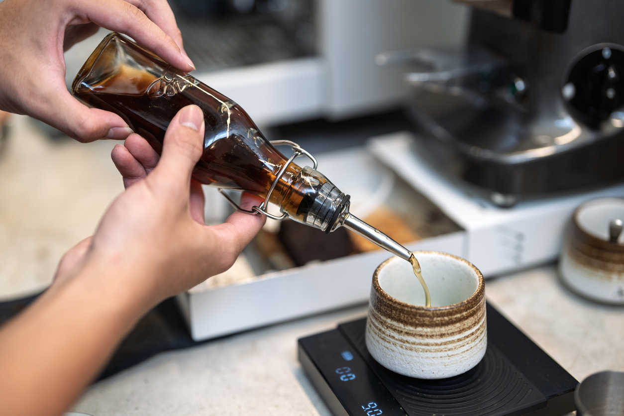 A person pours syrup from a glass bottle into a ceramic cup placed on a digital scale, with a coffee machine and other cups visible in the background.