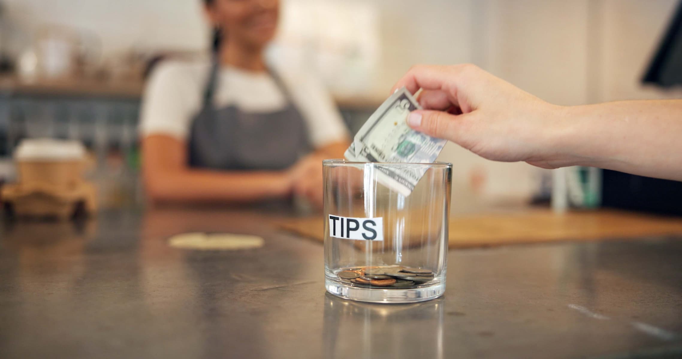 A hand places a dollar bill into a glass tip jar labeled "TIPS" on a counter, with coins inside. A person wearing an apron stands smiling in the blurred background.