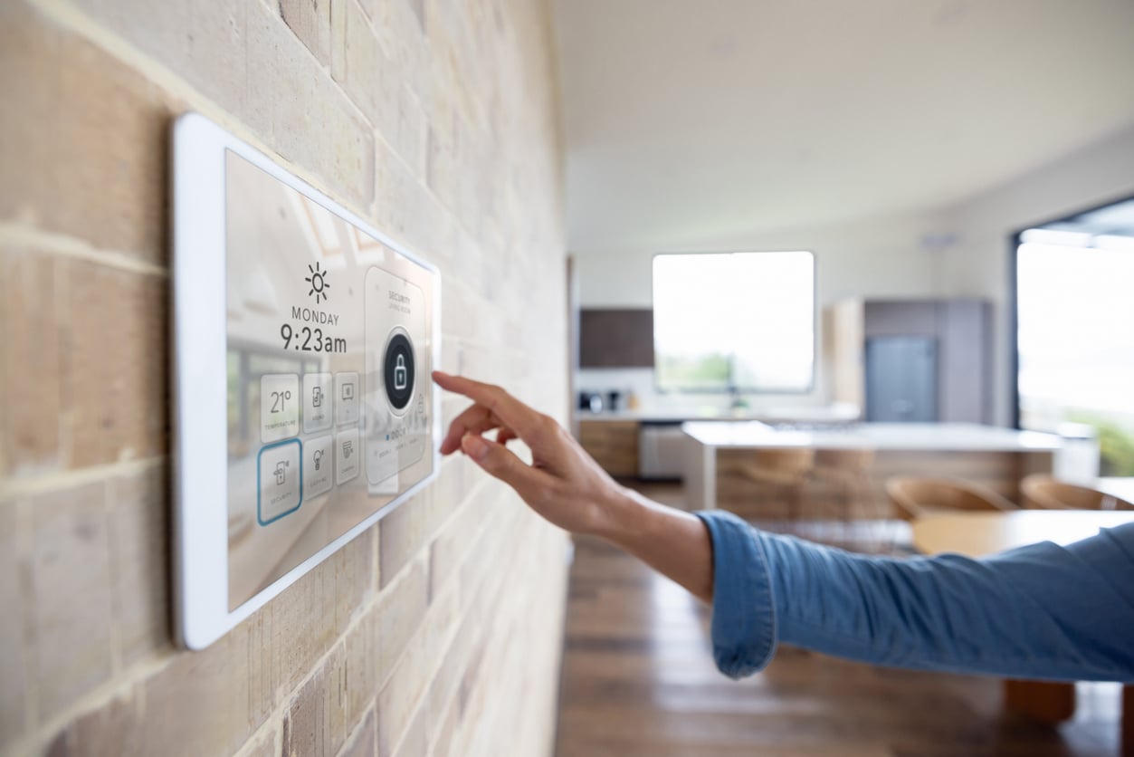 A person’s hand touches a wall-mounted smart home control panel displaying the time, date, weather, and security options in a modern, bright kitchen.
