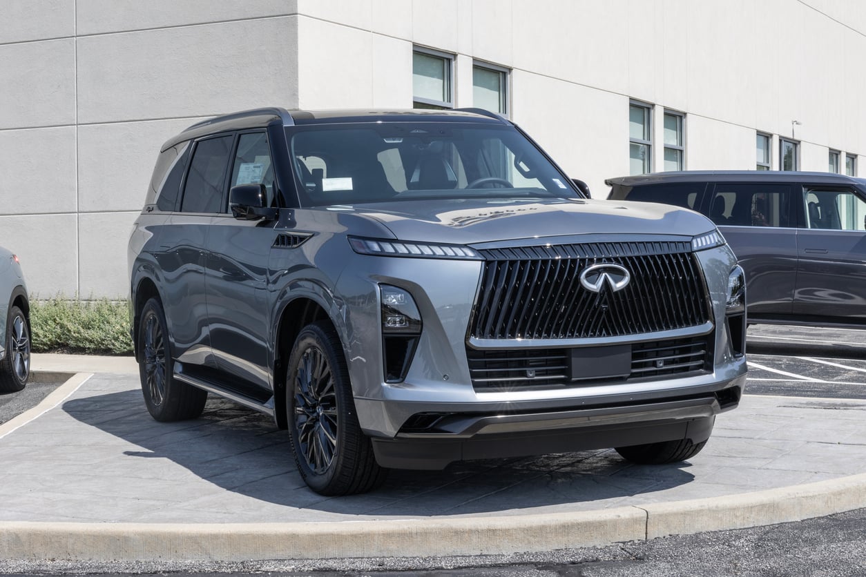 A silver Infiniti SUV is parked on a paved area near a modern building with large windows. The vehicle features a bold front grille and sleek, angular headlights.