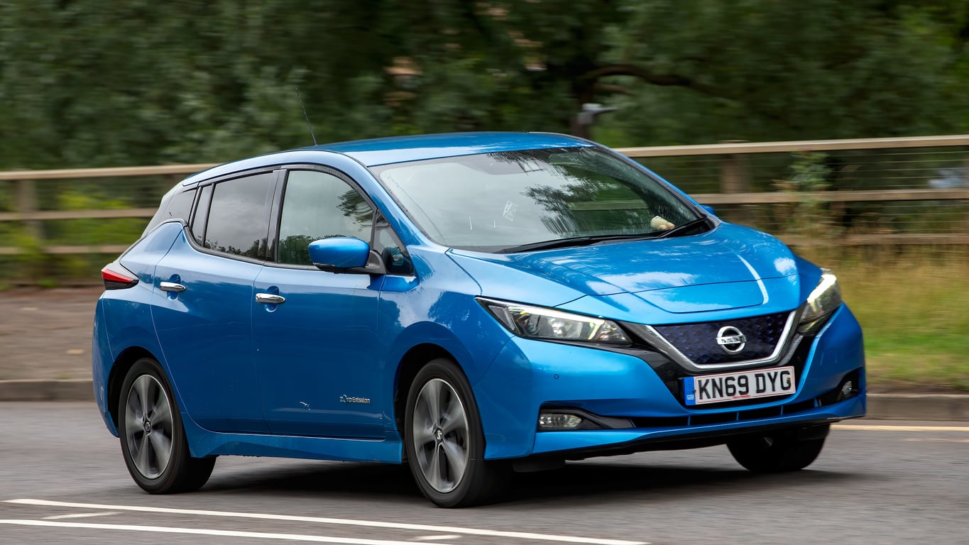 A blue Nissan Leaf electric car is driving on a road beside a wooden fence and green trees, with its headlights on and a UK license plate visible on the front.