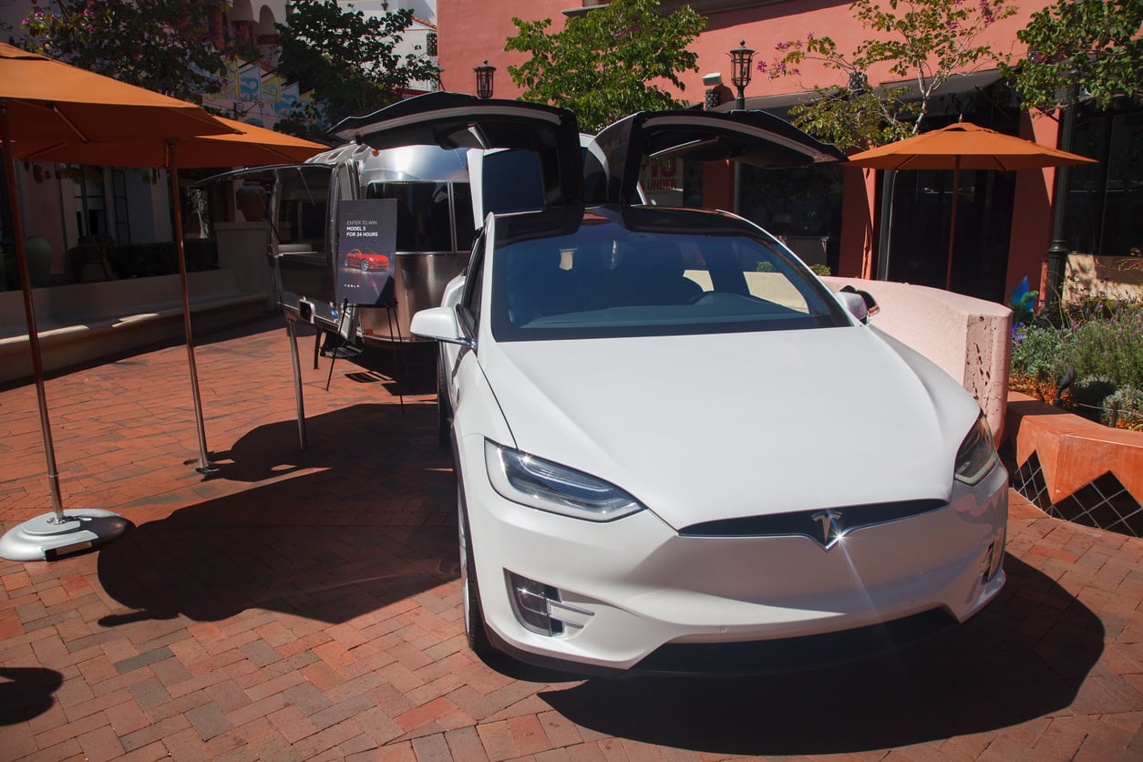 A white Tesla Model X with its falcon-wing doors open is parked on a brick patio, towing a silver Airstream trailer. Orange umbrellas provide shade nearby, and pink buildings and greenery are in the background.