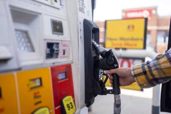 A person wearing a plaid shirt holds a gas pump nozzle at a fuel station, preparing to refuel a vehicle. The pump displays fuel grade options and signage is visible in the blurry background.
