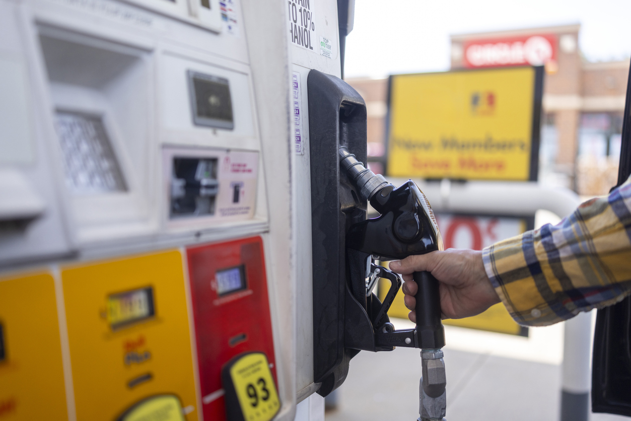 A person wearing a plaid shirt holds a gas pump nozzle at a fuel station, preparing to refuel a vehicle. The pump displays fuel grade options and signage is visible in the blurry background.