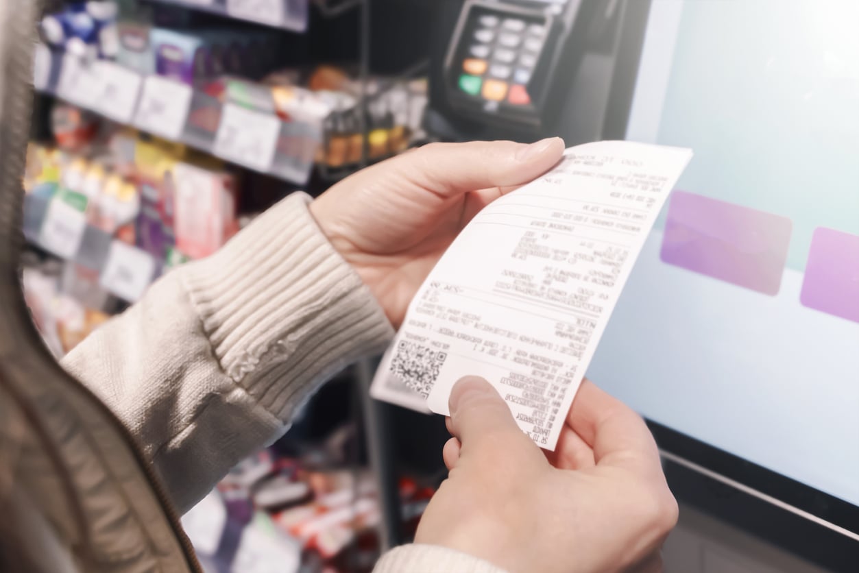 A person in a light sweater holds a printed receipt in front of a touchscreen machine at a store, with shelves of products and retail add-ons at checkout and a payment terminal visible in the background.