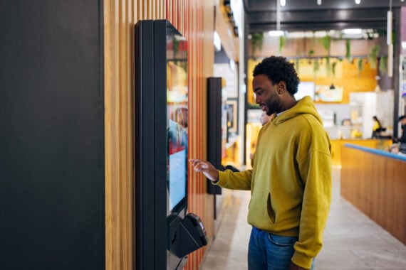 A man in a yellow hoodie uses a touchscreen ordering kiosk in a modern fast food restaurant. The interior features wood paneling, bright lighting, and a counter in the background.