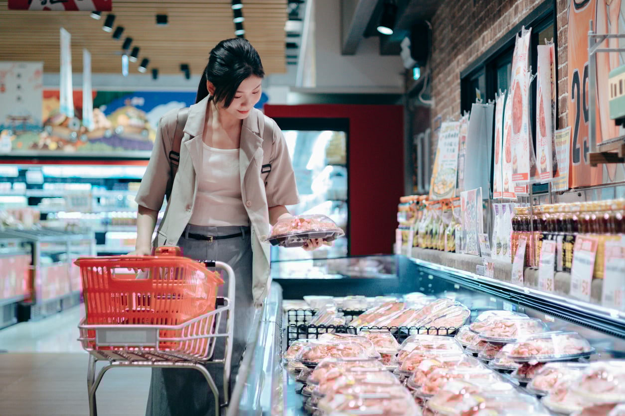 A woman shops in a grocery store, holding a basket in one hand while selecting packaged meat from a refrigerated display with the other hand. She is wearing a light blazer and appears focused on choosing her items.