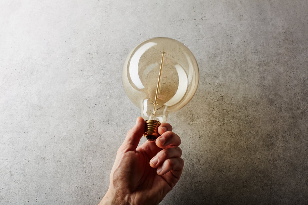 A hand holding a large, round, clear light bulb against a textured, light gray background. The light bulb is unlit and the filament inside is visible.
