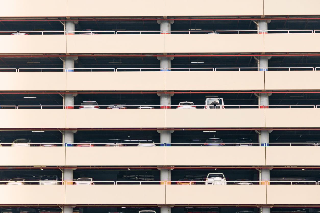 A multi-story parking garage with several parked cars visible on each level behind beige railings and columns. The image is taken from outside, showing a repetitive pattern of horizontal floors.