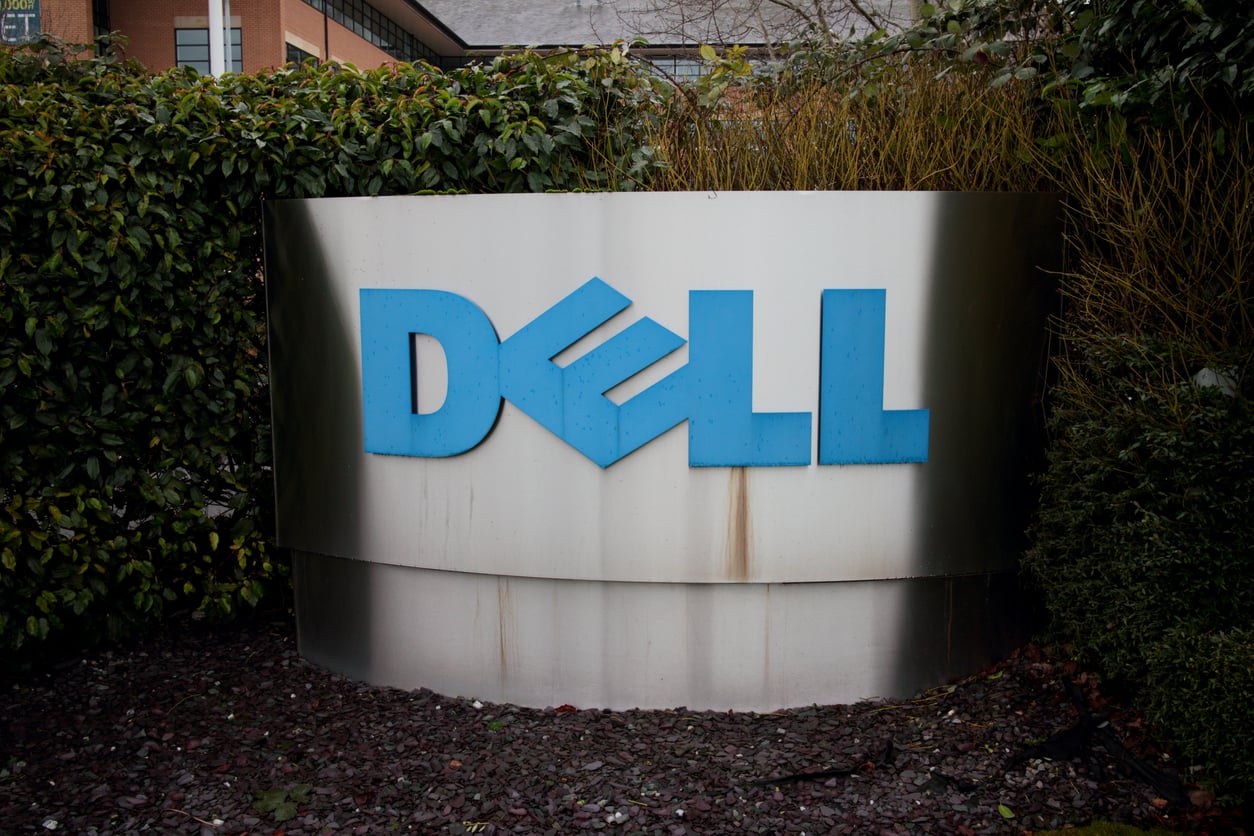 A large metallic sign with the blue Dell logo is displayed outdoors, surrounded by green bushes and plants, as discussions about the 2026 layoffs circulate within the company.