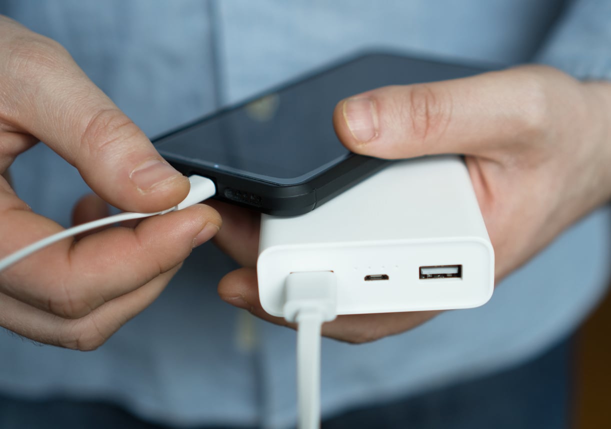 A person holds a smartphone in one hand and plugs a charging cable into it, while the other end is connected to a white power bank held in their other hand.
