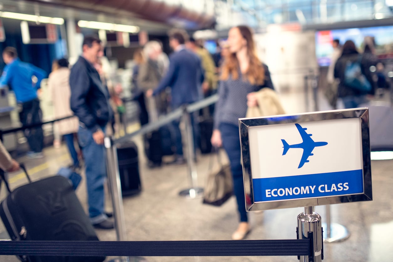 People stand in line at an airport check-in area, with a sign in the foreground reading "Economy Class" and showing an airplane icon. The scene appears busy, with travelers and luggage visible in the background.