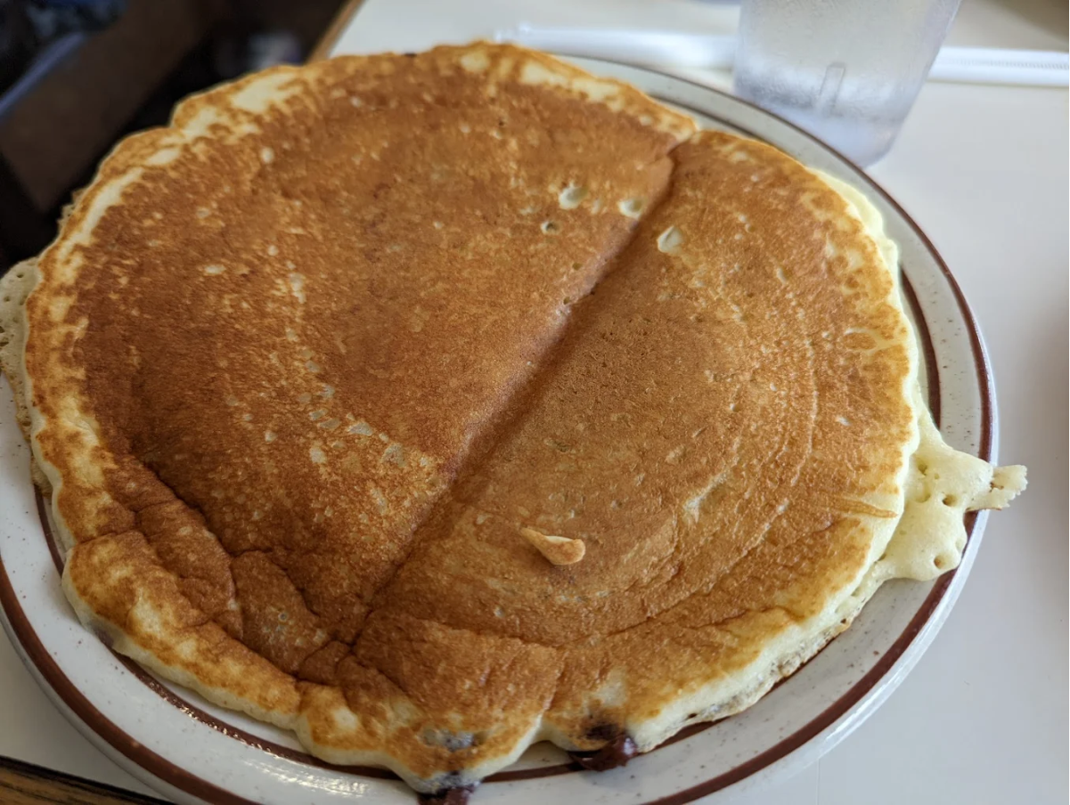 A large, golden-brown pancake on a white plate with a brown rim, sitting on a table next to a clear glass of water and a white straw.