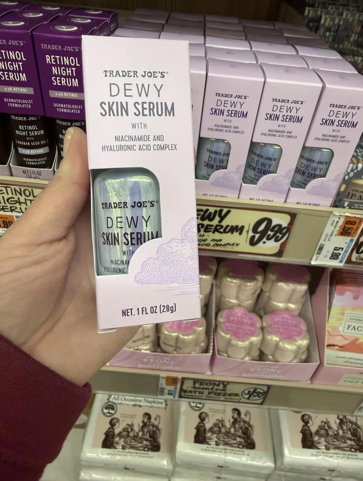 A hand holds a box of Trader Joe's Dewy Skin Serum with niacinamide and hyaluronic acid complex in front of a store shelf displaying more of the same product. Price tags and other skincare products are visible.