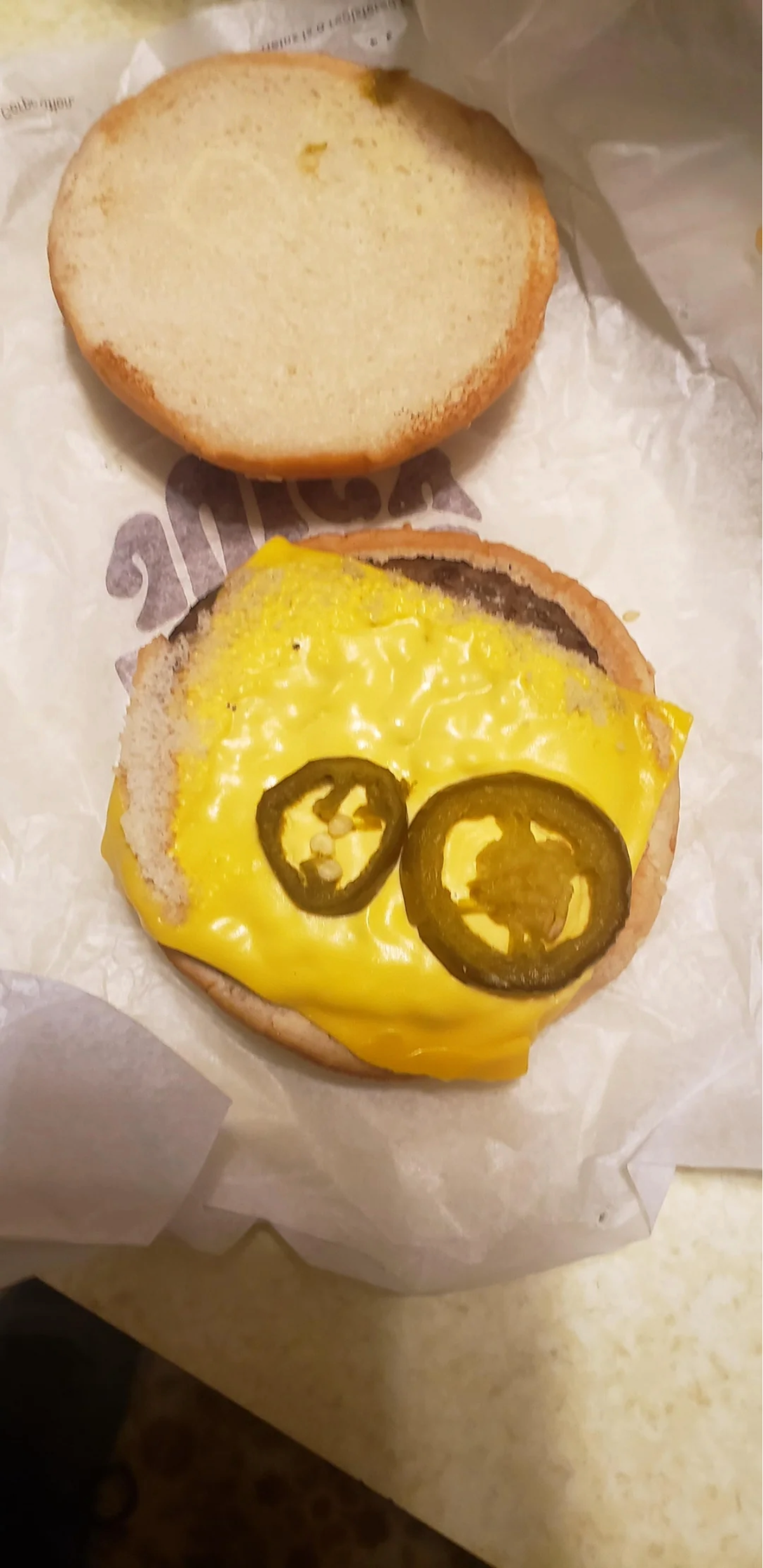 An open cheeseburger on wrapper paper shows a beef patty topped with melted yellow cheese and two jalapeño slices, with the top bun placed to the side.