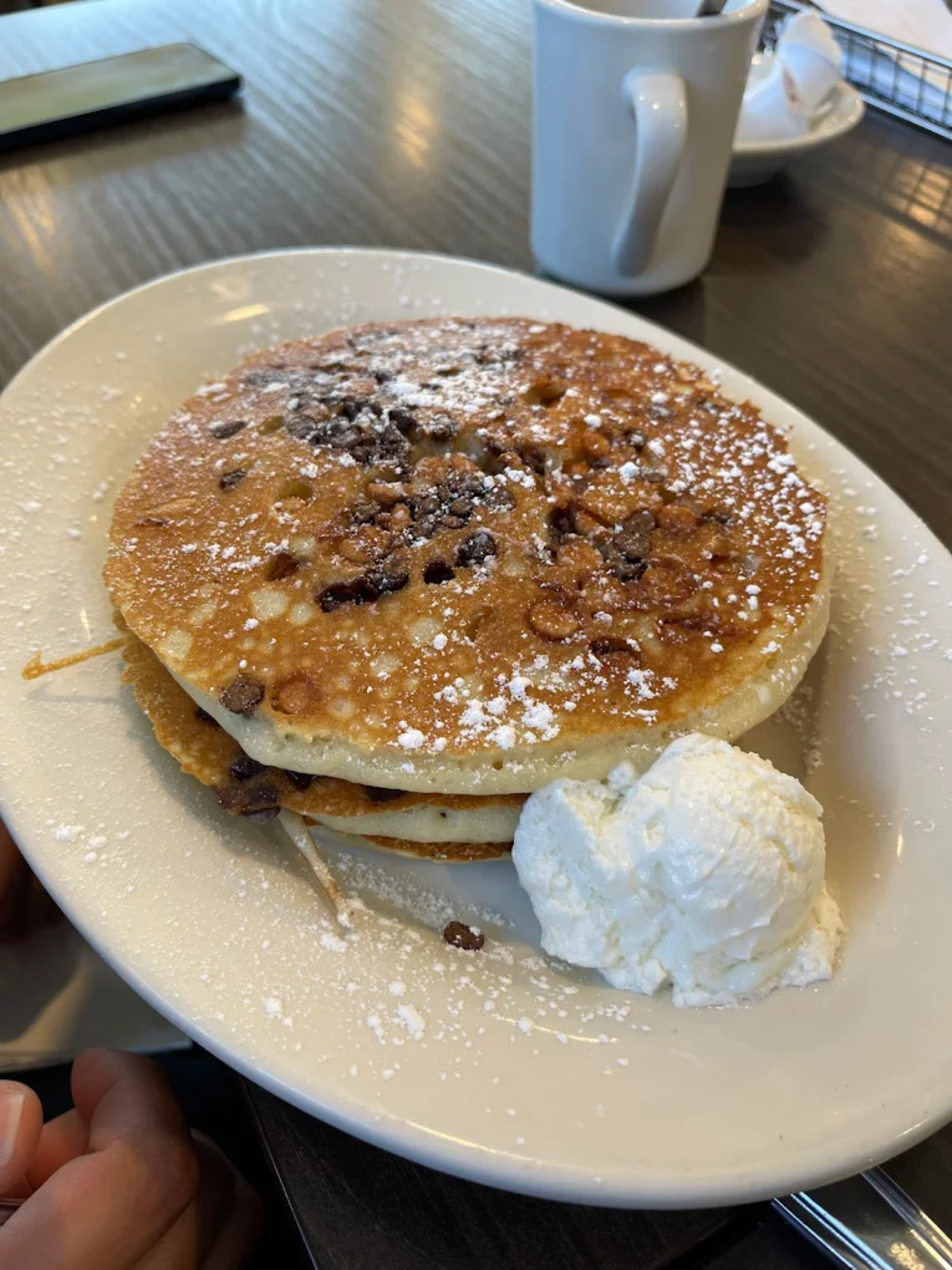A plate with two chocolate chip pancakes topped with powdered sugar, served with a scoop of whipped cream on the side. A white coffee mug and utensils are visible in the background.