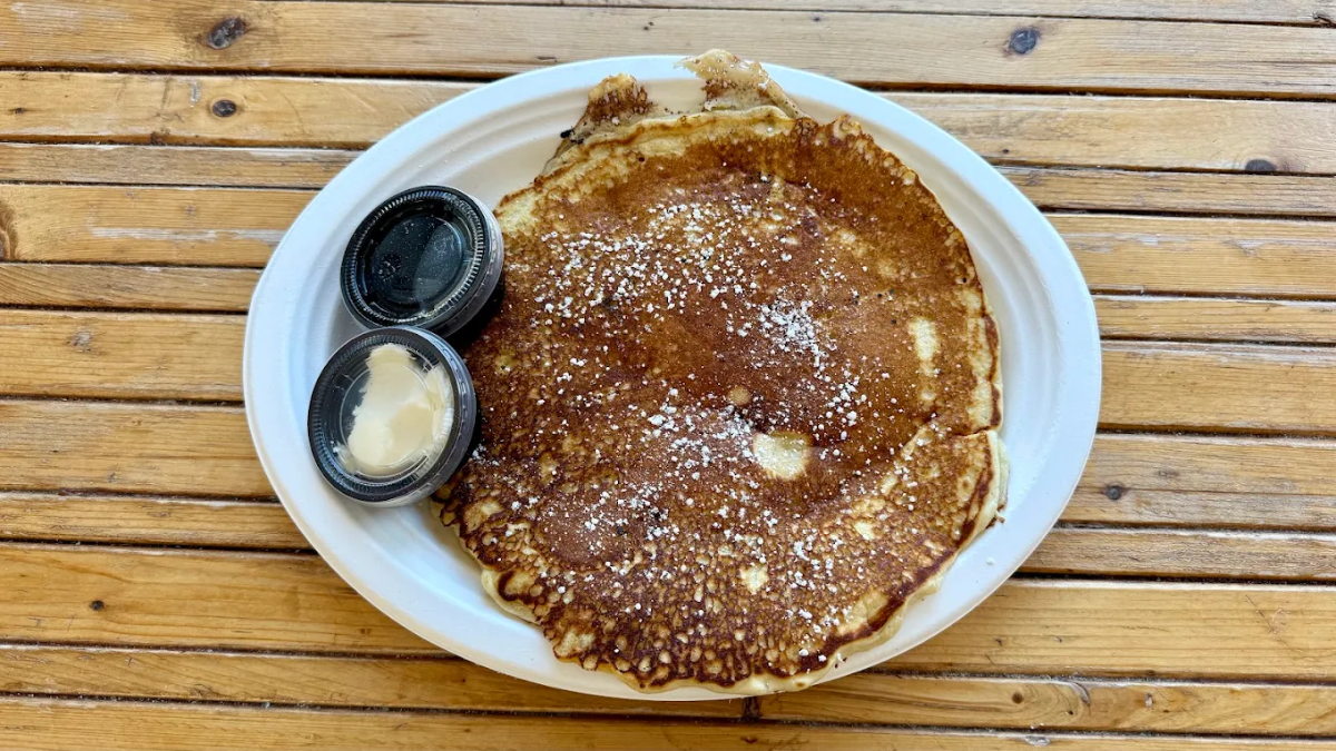A large pancake sprinkled with powdered sugar is served on a white plate with two small containers, one with butter and one with syrup, on a wooden table.