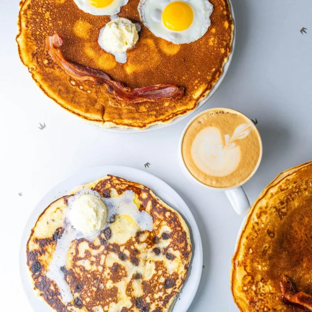 A breakfast scene with two plates of pancakes, one topped with eggs and bacon arranged as a smiley face, the other with melting butter, and a cup of latte with heart-shaped latte art on a white table.