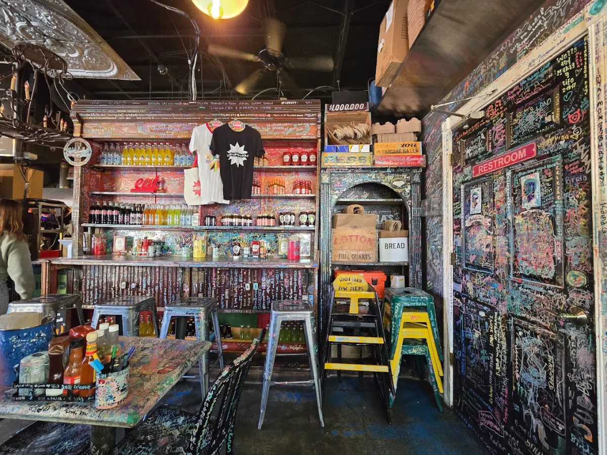 A vibrant, graffiti-covered bar interior with stools, shelves stocked with drinks, hanging t-shirts, and a door labeled "restroom." The walls and tables are covered in colorful writings and stickers.