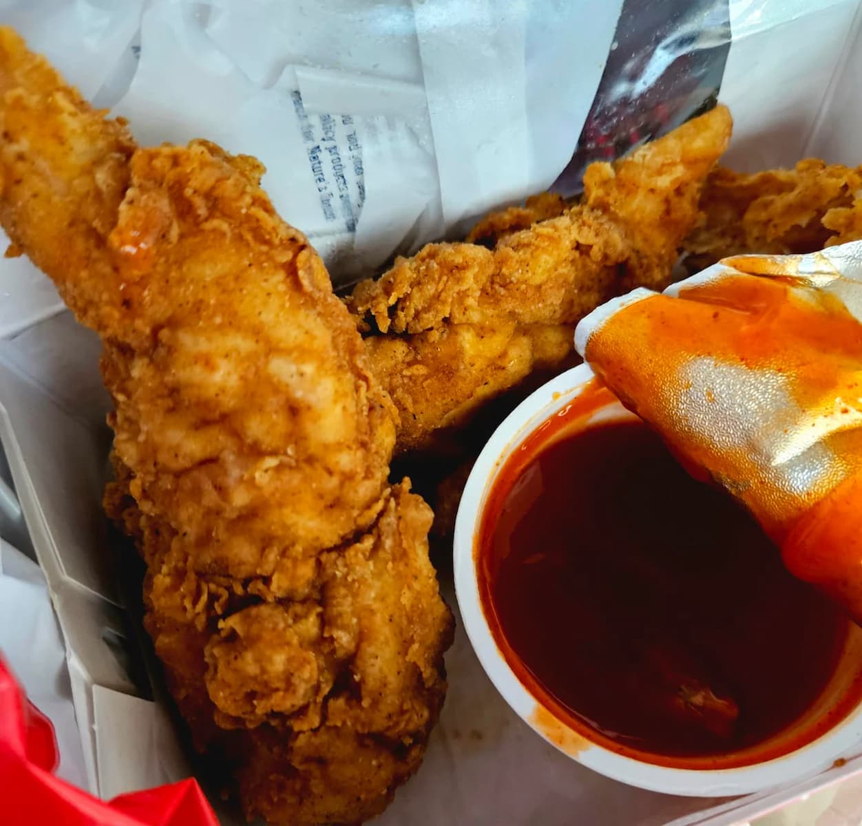 Close-up of crispy fried chicken tenders in a takeout box, with a container of red dipping sauce and a wrapper partially dipped in the sauce nearby.