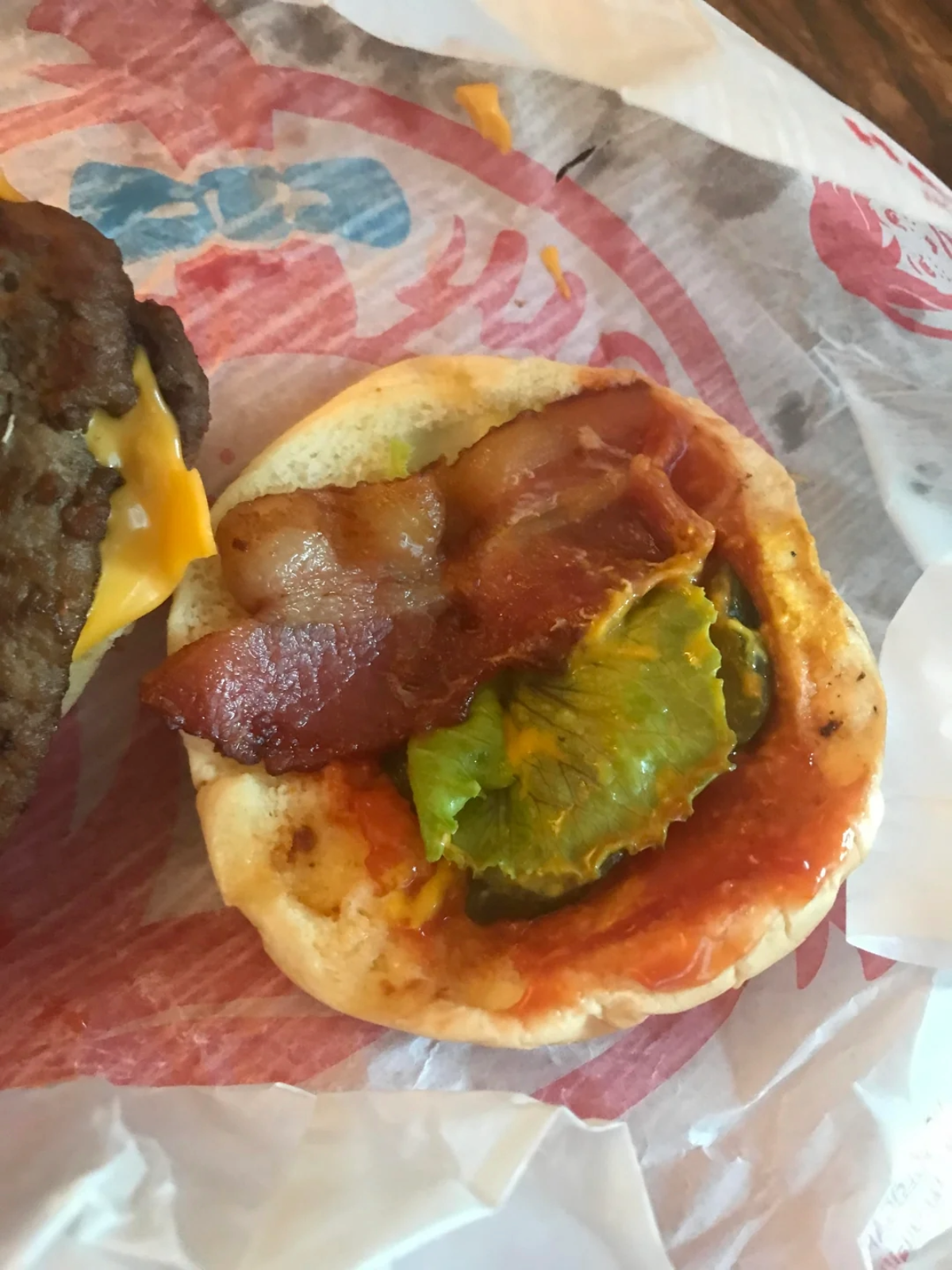 A hamburger bun topped with bacon, wilted lettuce, and ketchup sits open, with a slice of cheese and part of the burger patty visible on the side. The meal is on a fast-food wrapper.