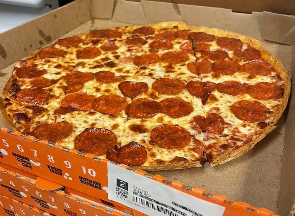 A pepperoni pizza with a thin crust sits in an open cardboard box, perfect for enjoying during march madness restaurant deals. The pizza is cut into slices and topped with melted cheese and pepperoni, while the box displays numbers and a barcode on the side.