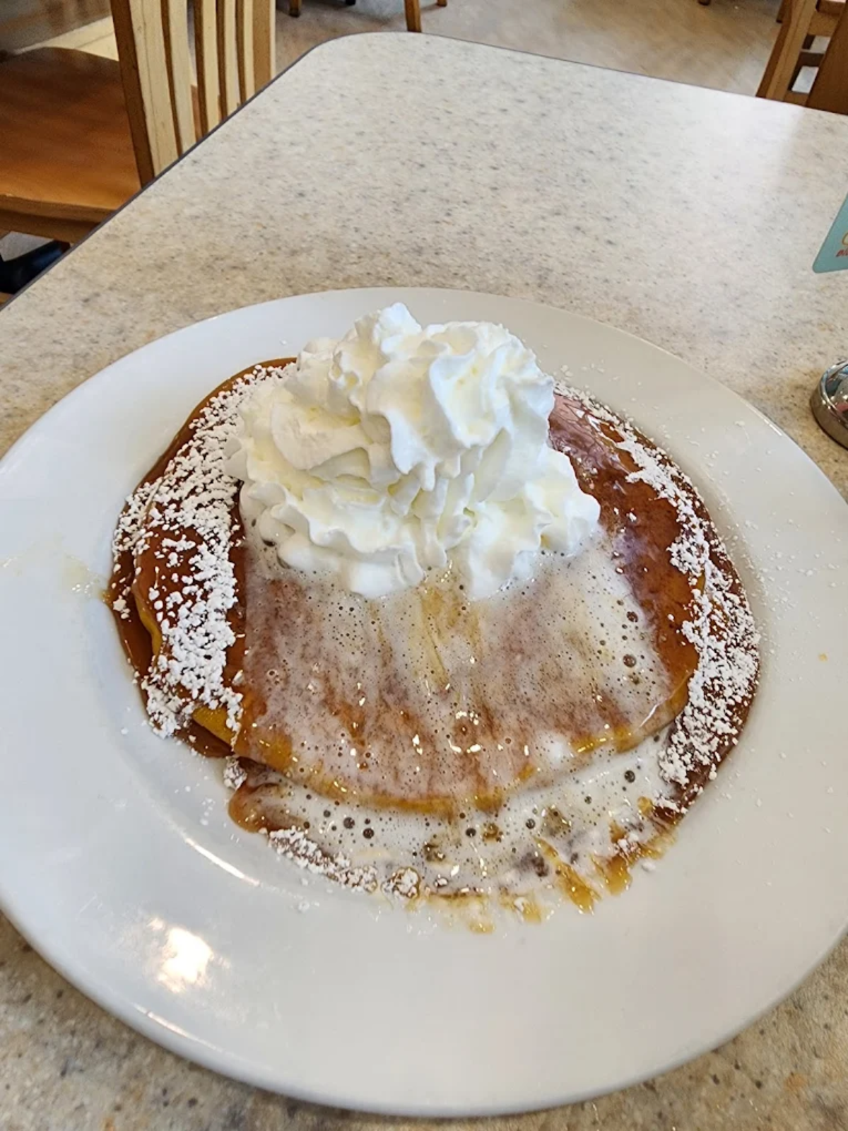 A plate with a large pancake topped with whipped cream, powdered sugar, and syrup, sitting on a light-colored table in a casual dining setting.