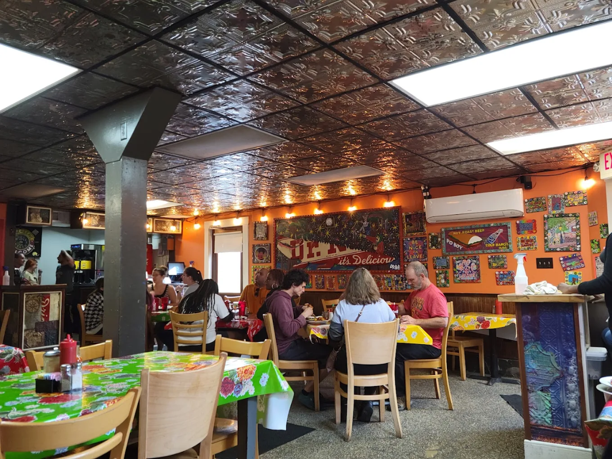 A colorful, lively restaurant interior with people dining at wooden tables. The walls are decorated with bright artwork and string lights. The ceiling has a metallic tile pattern, and there is a mix of patrons eating and talking.