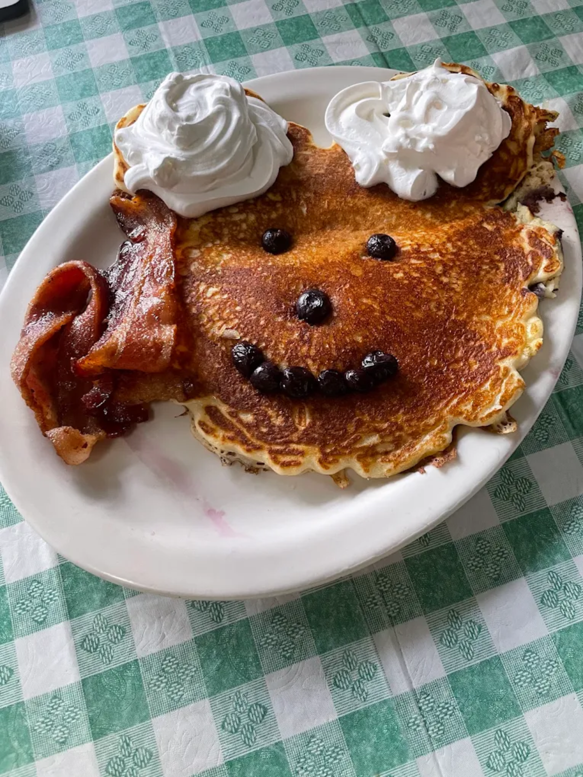 A pancake arranged to look like a smiling face, with whipped cream for hair, blueberries for eyes and a mouth, and two strips of bacon as ears, served on a white plate on a green checkered tablecloth.