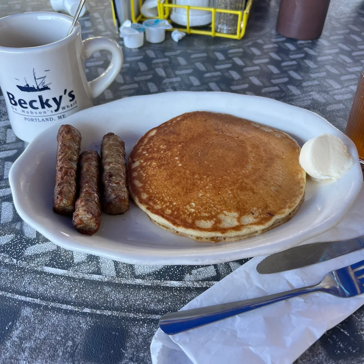 A breakfast plate with two sausages, a pancake with a pat of butter, a fork, a cup labeled "Becky's" with a spoon, and coffee creamers on a patterned table.