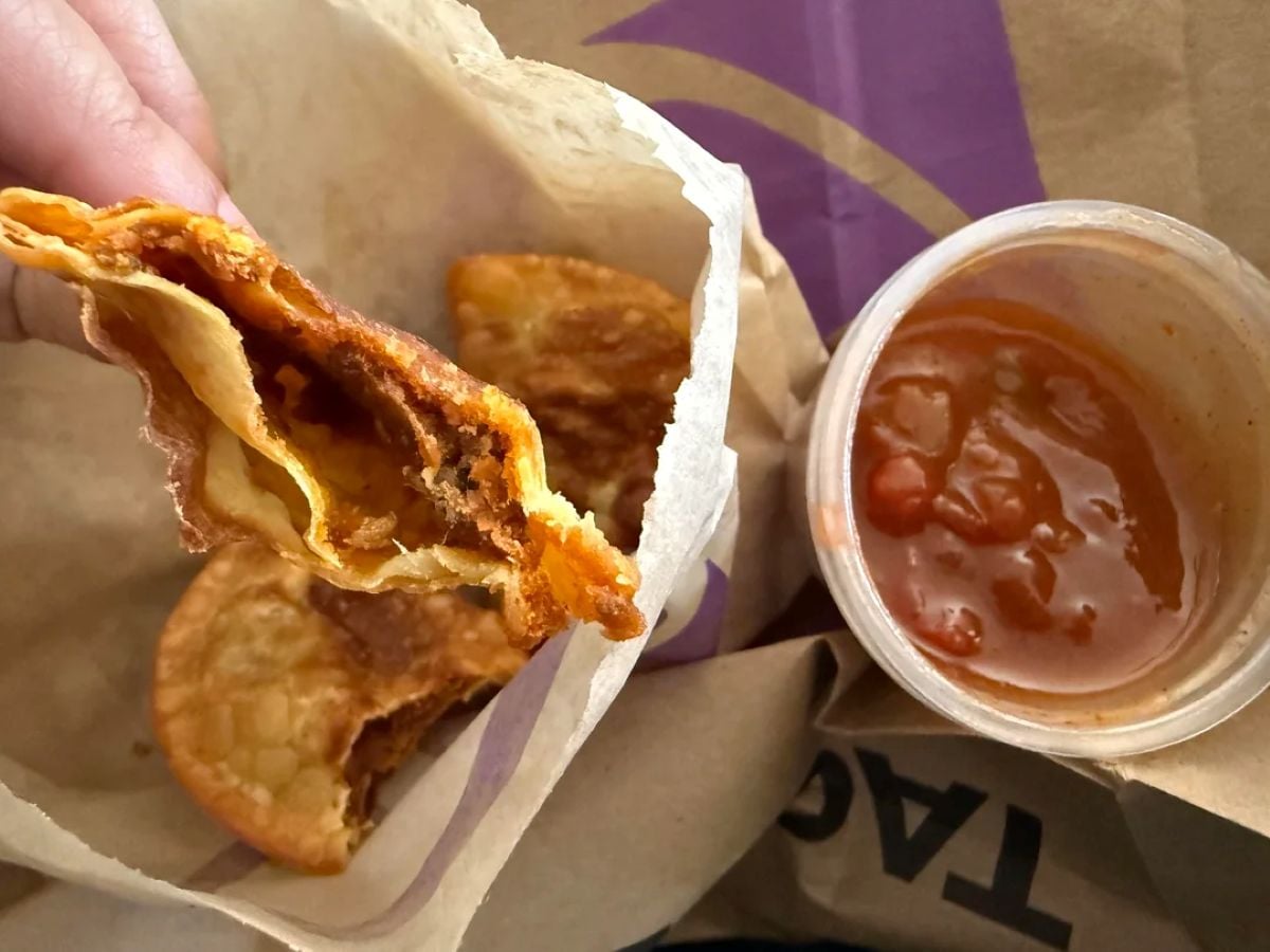 A hand holds a half-eaten Taco Bell quesadilla over a paper bag, with a small container of red salsa and two more quesadilla slices nearby.