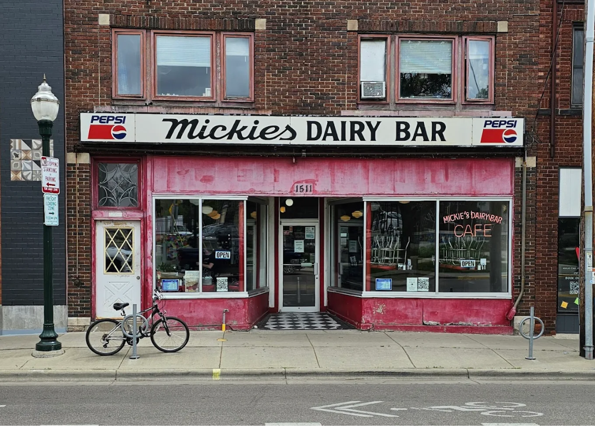 A brick building with a sign reading “Mickie’s Dairy Bar” above a red storefront. A bicycle is parked on the sidewalk, and the entrance has checkered flooring inside. Pepsi logos are displayed on the exterior sign.