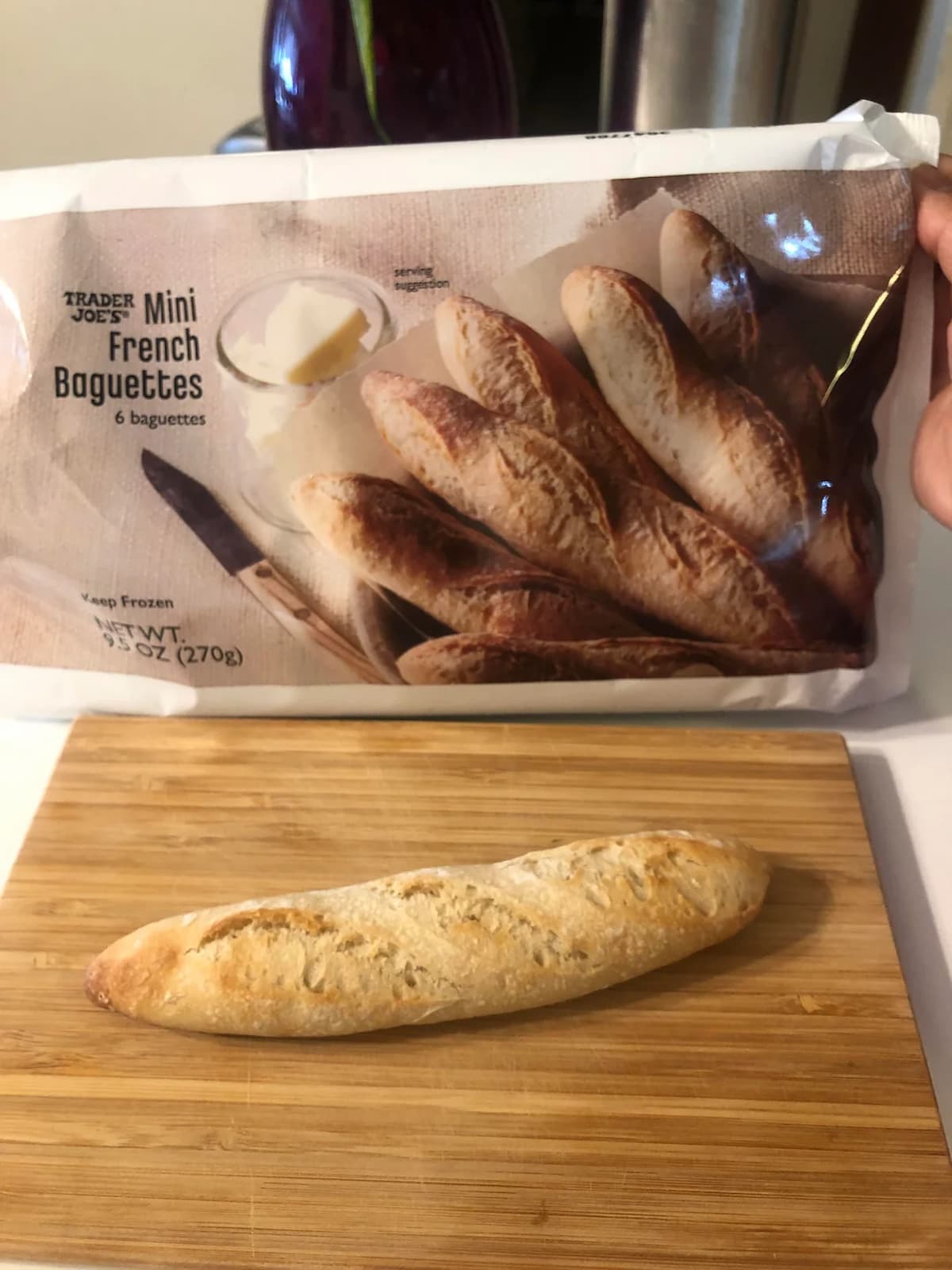 A single baked mini French baguette sits on a wooden cutting board in front of a Trader Joe's Mini French Baguettes package showing six baguettes, butter, and a knife.