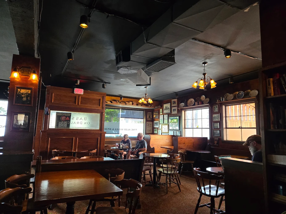 A cozy pub interior with wooden tables and chairs, warm lighting, framed pictures on the walls, and three people sitting at separate tables near large windows during daylight.