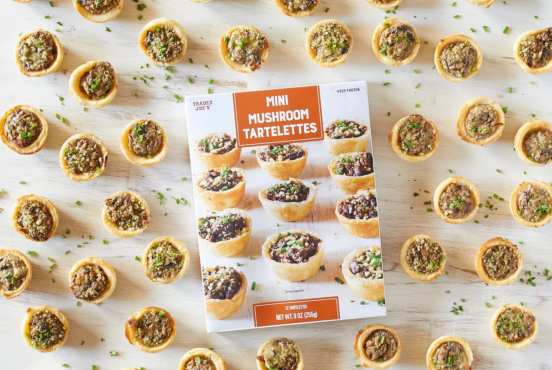 A box of Trader Joe’s Mini Mushroom Tartlets is surrounded by several cooked tartlets on a white surface, with chopped herbs scattered around.